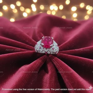 May include: A striking ring featuring a large oval ruby gemstone, surrounded by a halo of smaller diamonds and ruby accents. The ring is set on a silver band and displayed on a deep red velvet fabric. A luxurious piece of jewellery.