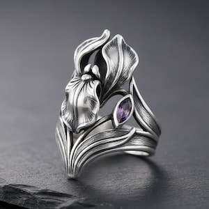 Puede incluir: Anillo de plata con diseño de flor de iris. El anillo presenta hojas y pétalos detallados, y una pequeña gema púrpura de corte ovalado. El anillo se encuentra sobre un fondo oscuro, resaltando el trabajo de metal.