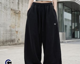 May include: Black wide-leg sweatpants with a drawstring waist and pockets. The pants have a small logo on the left leg. The image also shows a white crop top. The pants are part of a casual wear collection.