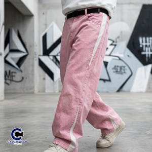 Peut inclure: Pantalon en jean rose clair avec une bande blanche sur le c&ocirc;t&eacute;. Le pantalon a une coupe ample et est port&eacute; avec une ceinture noire et des baskets blanches. L'arri&egrave;re-plan pr&eacute;sente du graffiti.