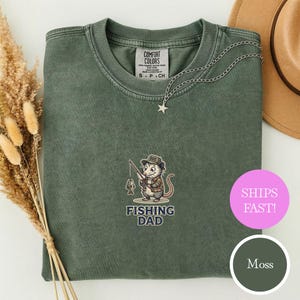以下が含まれることがあります： モスグリーンのTシャツ。帽子とジャケットを着た、釣りをするオポッサムのグラフィック。グラフィックの下には「FISHING DAD」の文字。星のペンダントが付いたシルバーのネックレスがシャツにかけられています。