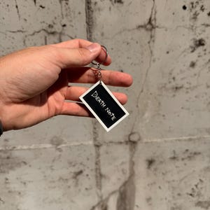 May include: A rectangular keyring featuring a black centre with the white text Death Note and a white border. The keyring is attached to a silver-coloured metal split ring by a short chain, providing a functional accessory for keys or bags.