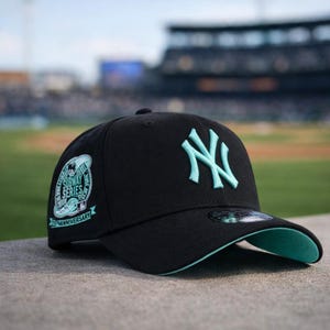 May include: Black baseball cap with a teal brim and the New York Yankees logo in teal. The cap features a Subway Series 25th Anniversary patch on the side. The cap is sitting on a concrete surface.