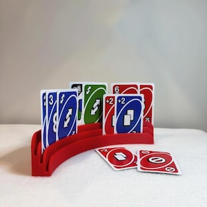 May include: A red card holder displaying a selection of Uno cards. The cards are blue, green, and red, featuring numbers and symbols. The holder is curved, designed to keep the cards upright.