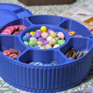 May include: A blue, circular game piece organiser with a lid, filled with various game components. The sections contain colourful tokens, wooden eggs, and coins. The organiser is made of plastic and is on a patterned surface. The diameter is approximately 15 cm.