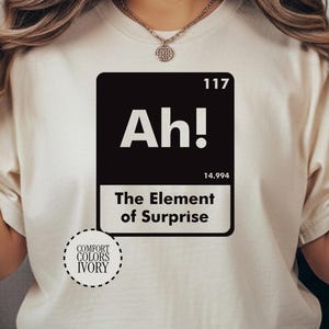 May include: Ivory-colored t-shirt with a black square graphic featuring the word "Ah!" and the phrase "The Element of Surprise." The graphic also includes the number "117" and the number "14.994." The shirt has a Comfort Colors Ivory label.