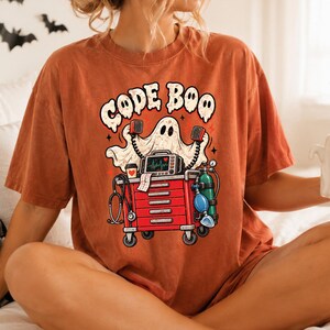 May include: A rust-colored t-shirt featuring a graphic of a ghost standing behind a red medical crash cart. The ghost holds two speakers, and the cart includes a heart monitor, stethoscope, and oxygen tank. The text "CODE BOO" is printed above.
