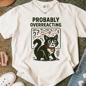 May include: An off-white short-sleeve t-shirt featuring the black text PROBABLY OVERREACTING above a graphic of a black and white cat with a worried expression, a red collar, and a background of red scribbled lines and question marks.