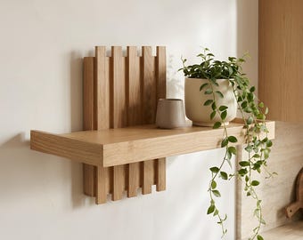 May include: A light-colored wooden wall shelf featuring a vertical slat design on the back panel. The shelf holds a small, smooth, light gray ceramic cup and a cream-colored ceramic planter containing a trailing green plant with small leaves.