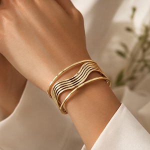 May include: A gold-toned cuff bracelet featuring a multi-layered design with five distinct, wavy, parallel bands that create a fluid, stacked appearance. The metal has a polished, reflective finish that catches the light across its curved surface.