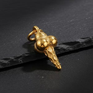 May include: A gold-colored pendant shaped like an ice cream cone with three scoops. The cone has a diamond pattern, and the pendant has a loop for a chain. The pendant is on a dark surface.