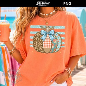 Op de afbeelding: Een perzikkleurig t-shirt met een afbeelding van een pompoen met een lichtblauwe strik. De pompoen heeft secties met luipaardprint, oranje-witte ruitjes en bloemmotieven, geplaatst tegen een achtergrond van horizontale lichtblauwe strepen.
