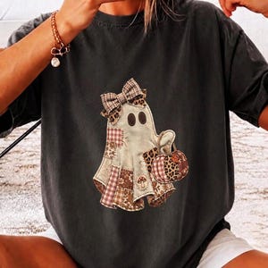 以下が含まれることがあります： チャコールグレーの半袖Tシャツ。中央にはチェック柄のリボンをつけたゴーストのグラフィックが描かれています。ゴーストは茶色のチェック柄、ヒョウ柄、花柄などの様々なパッチワーク模様で構成されており、カボチャを持っています。