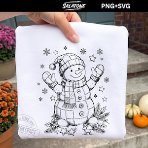 Könnte beinhalten: Ein weißes T-Shirt mit einer schwarzen Strichzeichnung eines Schneemanns, der eine Mütze, einen Schal und Fäustlinge trägt. Der Schneemann ist von Schneeflocken, Sternen und Stechpalmenblättern umgeben. Unten ist ein kleines Rentier abgebildet.