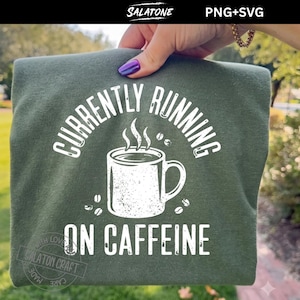 May include: An olive green t-shirt featuring a white graphic of a coffee mug with steam rising from it. The text "CURRENTLY RUNNING ON CAFFEINE" is printed in a white arched font above and below the mug, accompanied by small coffee bean icons.