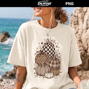 May include: A cream-colored short-sleeve t-shirt featuring a graphic print of five pumpkins with various patterns including leopard print, plaid, and checkered designs, accented with small stars and leaves in shades of brown and black.