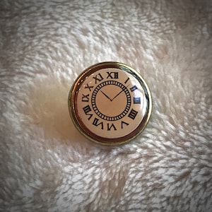 May include: A gold-colored pin with a cream-colored clock face. The clock face has black Roman numerals and a black hour and minute hand. The pin is on a white fuzzy surface.