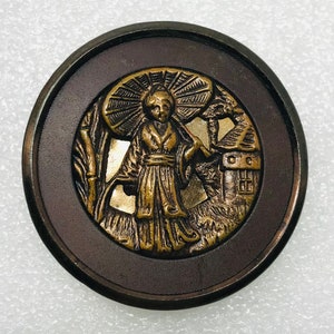May include: A round, antique button with a detailed relief of a woman in traditional Japanese attire holding an umbrella. The central image is gold-toned, set against a brown background and a dark brown outer ring. The scene includes bamboo and a small building.
