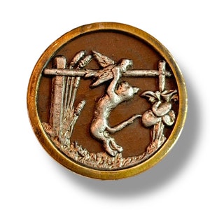 May include: A round, brass-colored button with a raised, silver-colored design. The design depicts a cat jumping over a fence with a bird in its mouth. The background is brown with a fence and grass.