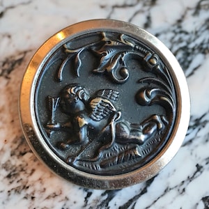 May include: A silver and black metal button with a raised design of a winged cherub holding an arrow. The cherub is surrounded by a floral design.