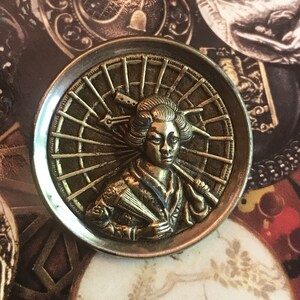 May include: A brass button with a raised design of a woman holding an umbrella. The woman is wearing a traditional Japanese kimono and has her hair styled in a bun. The umbrella is open and has a detailed design.