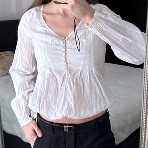 May include: A white long-sleeved blouse featuring a V-neckline, a central vertical row of small buttons, and a gathered, elasticized hemline. The fabric has a textured, slightly crinkled appearance, and the sleeves are loose with fitted cuffs.