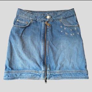 May include: Light blue denim skirt with a front zipper closure and a small pocket on the left side. The skirt features embroidered white star accents near the right pocket. The skirt has a button closure at the waistband.