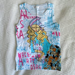 May include: White vest top with a colourful graphic print. The design features a cartoon illustration of a woman with long blonde hair, wearing a blue top and floral skirt, holding a phone. The background has text in pink and blue.