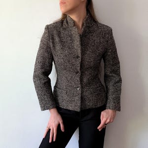 May include: A brown and black herringbone patterned blazer featuring a structured collar, long sleeves, and a three-button front closure. The jacket is tailored with a fitted silhouette and includes two horizontal flap pockets at the waistline.