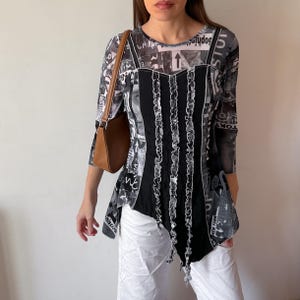 May include: A long-sleeve top featuring a black and white collage print with text including Paris and New York, a solid black central panel with vertical ruffled trim, and a brown shoulder bag paired with white trousers.