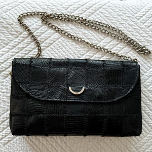 Peut inclure: Un sac à bandoulière rectangulaire noir présentant un motif texturé en forme de grille sur toute la surface. Le sac possède un rabat incurvé avec un accent métallique semi-circulaire argenté et une longue bandoulière en chaîne métallique argentée.