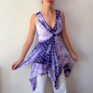 May include: A sleeveless tunic top featuring a deep V-neckline and a handkerchief hemline. The garment displays a purple and white tie-dye pattern with circular motifs radiating from the waist, paired with plain white trousers.