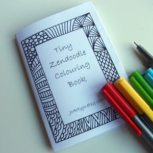 May include: A small, white book with a black and white patterned border. The title of the book is "Tiny Zendoodle Colouring Book". The book is lying on a white surface with five colored pens next to it.