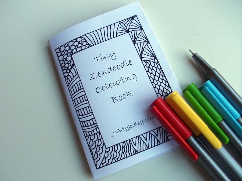 May include: A small, white book with a black and white patterned border. The title of the book is "Tiny Zendoodle Colouring Book". The book is lying on a white surface with five colored pens next to it.