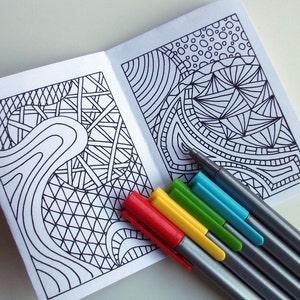 May include: A small, blank coloring book with two pages of intricate black and white patterns. The book is open and lying flat on a white surface. Six multi-colored markers are lying on top of the book.