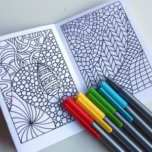 May include: A small, blank coloring book with two pages of intricate black and white patterns.  The book is open and lying on a white surface.  A set of seven colored markers are lying on top of the book.