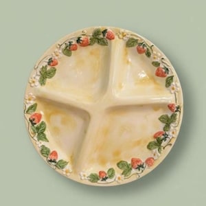 May include: A round, divided serving dish with a cream-colored interior and a decorative strawberry and floral border. The dish is divided into four sections and features red strawberries, green leaves, and white flowers around the rim.
