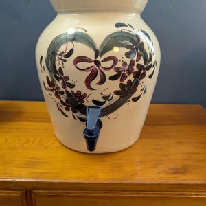 May include: Cream-colored ceramic water dispenser with a heart-shaped floral design in black, green, and burgundy. Features a blue spigot. The dispenser sits on a wooden surface.
