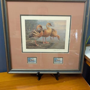 May include: A framed print featuring three brown ducks standing on a log in a misty wetland. The artwork includes a tan mat, a dark frame, and two small postage stamps depicting the same duck scene mounted below the main illustration.