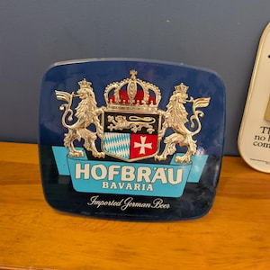 May include: A blue Hofbrau Bavaria sign with a crest featuring lions, a crown, and a shield with a cross. The sign also includes the text "HOFBRÄU" and "Imported German Beer."
