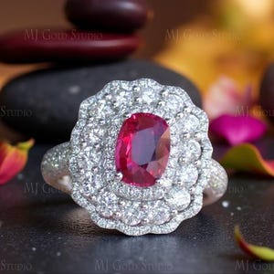 Op de afbeelding: Een zilveren ring met een grote, ovaal geslepen robijn als middensteen, omringd door een halo van kleinere, sprankelende diamanten. Het ontwerp van de ring heeft een bloemachtig geïnspireerde zetting, met ingewikkelde details op de band. De achtergrond is wazig, met de tekst "MJ Gold Studio".