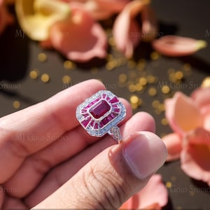 May include: A silver ring featuring a rectangular ruby centre stone, encircled by smaller rubies and clear accent stones. The ring has an Art Deco design, with the text "MJ Gold Studio" visible in the background.