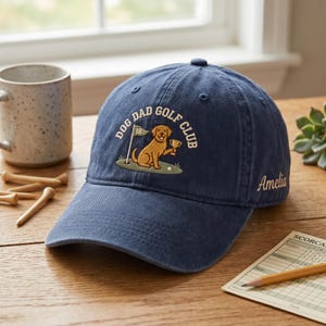May include: A navy blue baseball cap featuring an embroidered graphic of a golden retriever holding a golf trophy on a green with a flag labeled 18 and the text DOG DAD GOLF CLUB. The name Amelia is embroidered in white on the side of the cap.