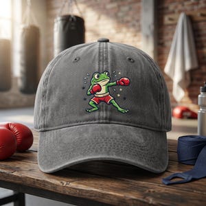 May include: Gray baseball cap featuring a cartoon frog in boxing attire. The frog is green with red boxing gloves and shorts, striking a boxing pose. The cap is made of a textured fabric and has a curved brim.