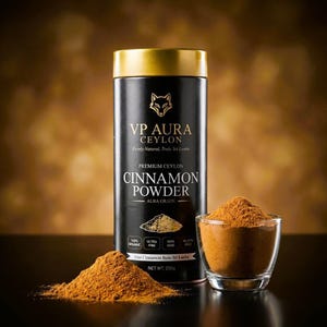 Op de afbeelding: Een zwarte en gouden cilindrische container met VP Aura Ceylon kaneelpoeder, met een voslogo. De afbeelding toont ook een klein glas kaneelpoeder en een hoop kaneelpoeder. De tekst op de container luidt "Premium Ceylon Cinnamon Powder, Alba Grade".