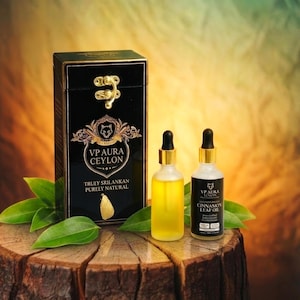 Könnte beinhalten: Eine schwarze Schachtel mit goldenen Verzierungen und einem goldenen Verschluss, beschriftet mit "VP Aura Ceylon", steht neben zwei Glasflaschen mit Öl. Eine Flasche ist klar mit gelber Flüssigkeit, die andere ist schwarz mit weißem Text. Grüne Blätter umgeben die Produkte.
