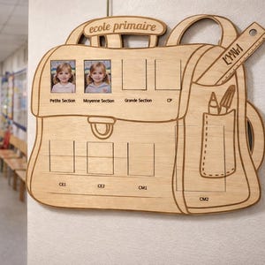 Peut inclure: Panneau en bois en forme de sac à dos scolaire avec des détails découpés. Il comporte des espaces pour les photos et les niveaux scolaires, avec les mots "ecole primaire" en haut. Une règle avec le nom "LYNN" est attachée sur le côté.