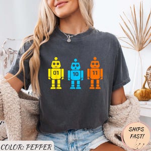 Puede incluir: Camiseta gris oscuro con tres gráficos de robots coloridos. Cada robot es amarillo, azul o naranja y tiene los números 01, 10 y 11 en el pecho. El texto "COLOR: PEPPER" está en la parte inferior.