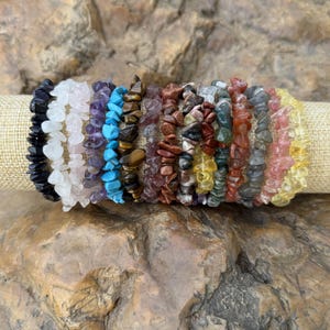 May include: A collection of gemstone chip bracelets in various colors, including black, white, purple, turquoise, brown, red, yellow, and pink. The bracelets are displayed on a beige fabric roll against a natural stone background.