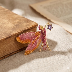 May include: A gold-coloured brooch depicting a fairy holding a purple star. The fairy has pink and orange wings and a pink dress. The brooch is set against a neutral background with a vintage book.